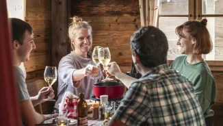 Two women and two men sit together at a table in a wooden chalet and enjoy a cheese fondue. They toast with a glass of white wine.