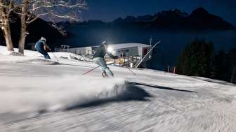 Two skiers look out over the illuminated Rhine Valley while skiing at night on the Pizol