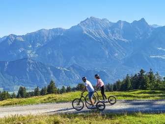 Two people ride downhill on Alpine scooters and the mountains of the Bündner Herrschaft can be seen in the background