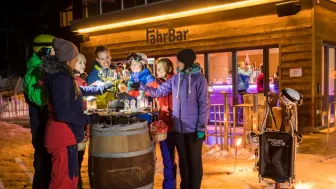 A group of winter sports enthusiasts stand in front of the snow-covered FahrBar and toast with glasses of wine.