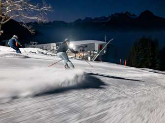Two skiers look out over the illuminated Rhine Valley while skiing at night on the Pizol